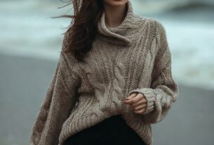 Stunning young woman walking on a grey windy beach wearing a chunky knit sweater and tailored shorts, moody coastal light, photoreal 8K
