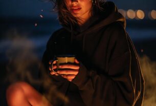 Stunning young woman sitting on a driftwood log at a beach bonfire at night wearing an oversized hoodie and high-waisted bikini bottoms, sparks in the air, photoreal 8K
