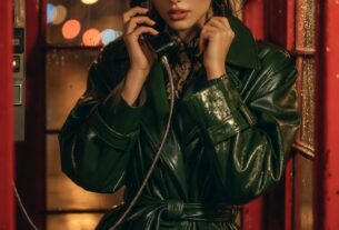 Stunning young woman inside a London telephone booth talking on the phone with rain on the glass, wearing a glossy mac coat, cinematic streetlight bokeh, photoreal 8K