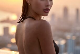 Young European woman leaning on a rooftop infinity pool edge at sunset with city skyline reflections and wet slicked-back hair