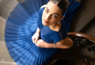 Stunning young woman on a spiral staircase looking up, photographed from above in a flowing pleated dress that spirals around her, photoreal 8K