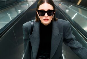 Young European woman looking up at the camera on an escalator wearing oversized sunglasses and a chic coat in a cold metallic urban setting