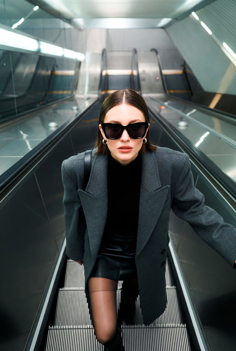 Young European woman looking up at the camera on an escalator wearing oversized sunglasses and a chic coat in a cold metallic urban setting