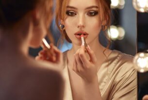 Young European redhead applying lipstick while looking into a Hollywood-style lighted vanity mirror, silk robe slipping off one shoulder