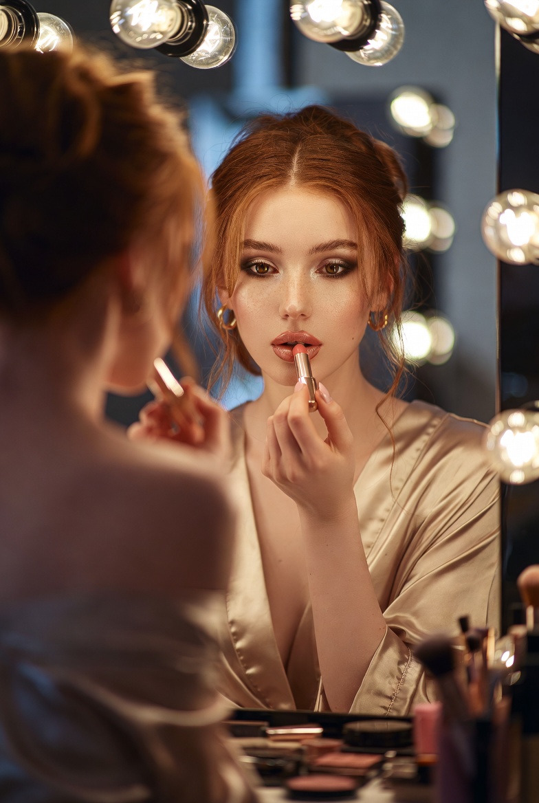 Young European redhead applying lipstick while looking into a Hollywood-style lighted vanity mirror, silk robe slipping off one shoulder