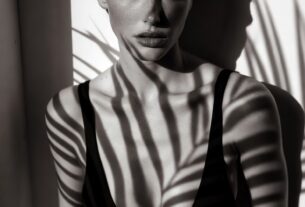 Young European woman in a minimalist black swimsuit standing against a white wall with palm-leaf shadows across her face in high-contrast black and white